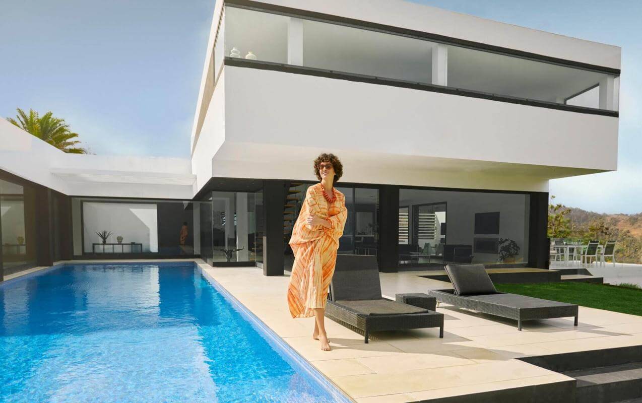 Persona a bordo piscina privata di fronte a villa moderna dalle linee minimaliste, con lettini e cielo limpido.