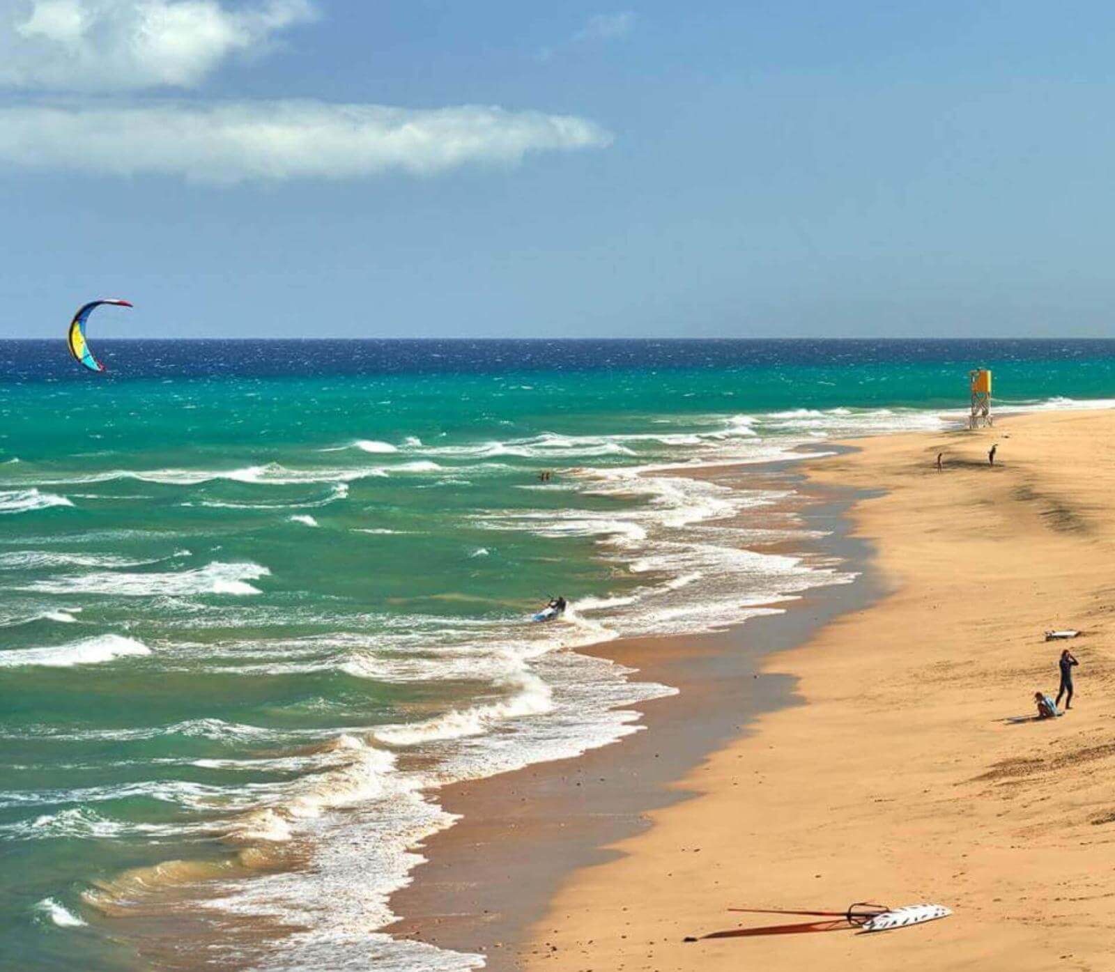 Spiaggia di sabbia dorata con onde e persone che praticano sport acquatici come il kitesurf.