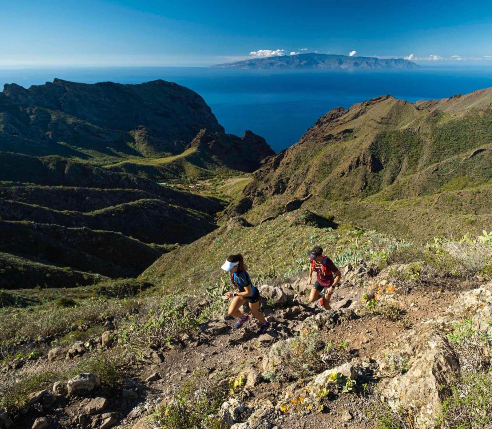 Due persone che praticano trail running su sentieri montuosi con vista sull'oceano e un'altra isola all'orizzonte.