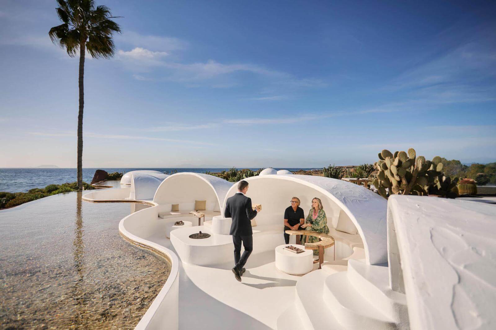 Persone che conversano su una terrazza bianca con design organico vicino alla piscina e vista sul mare.
