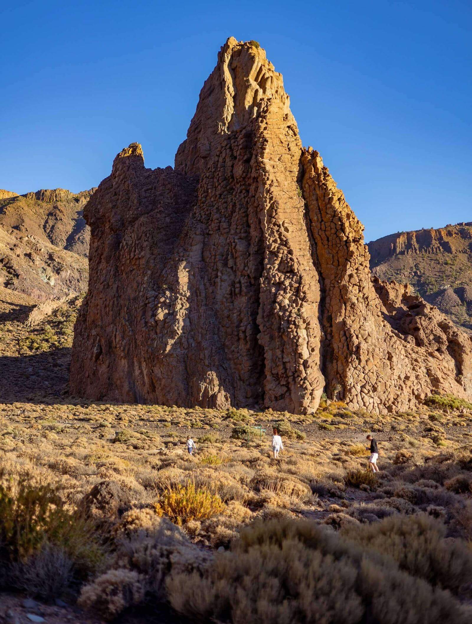 Gente che cammina tra i cespugli davanti a una grande formazione rocciosa verticale in un paesaggio vulcanico.