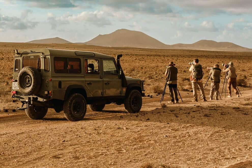 Gruppo con fotocamere e treppiedi che osserva gli uccelli in un paesaggio arido accanto a un fuoristrada.
