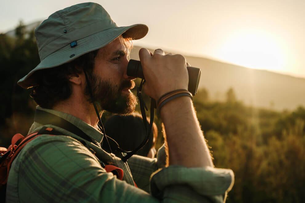 Primo piano di una persona con un cappello da trekking che osserva il paesaggio con un binocolo al tramonto.