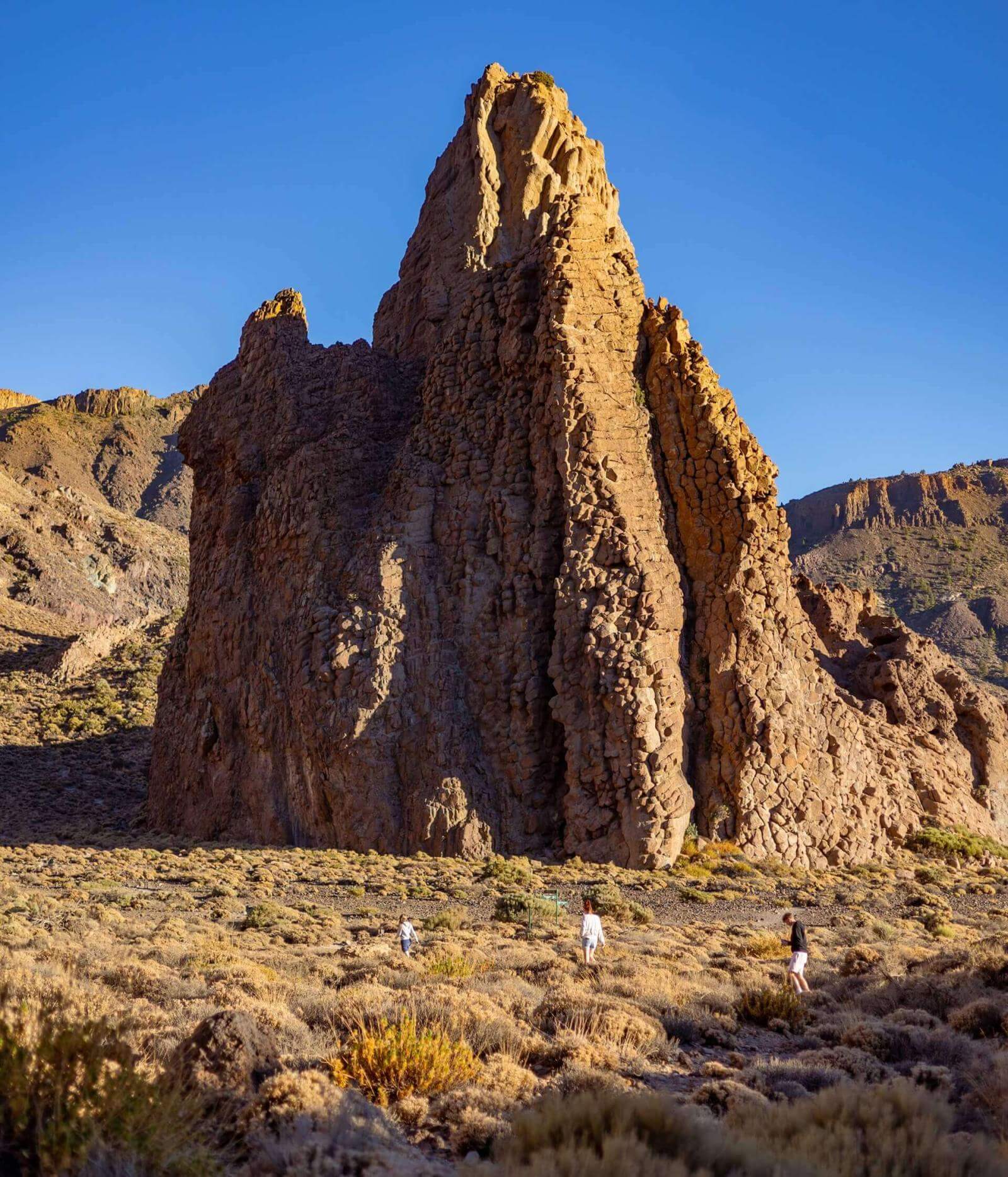 Gente che cammina tra i cespugli davanti a una grande formazione rocciosa verticale in un paesaggio vulcanico.