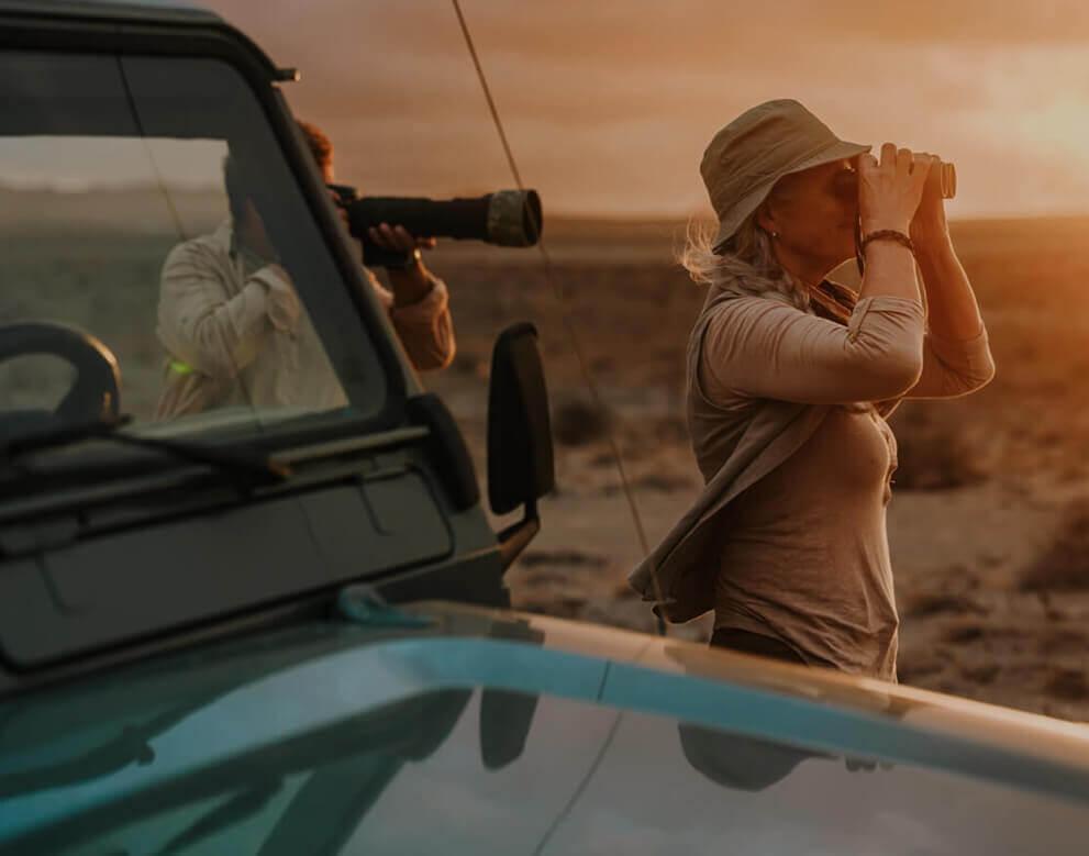 Persona che osserva gli uccelli con il binocolo in un paesaggio arido al tramonto, accanto a un fuoristrada.