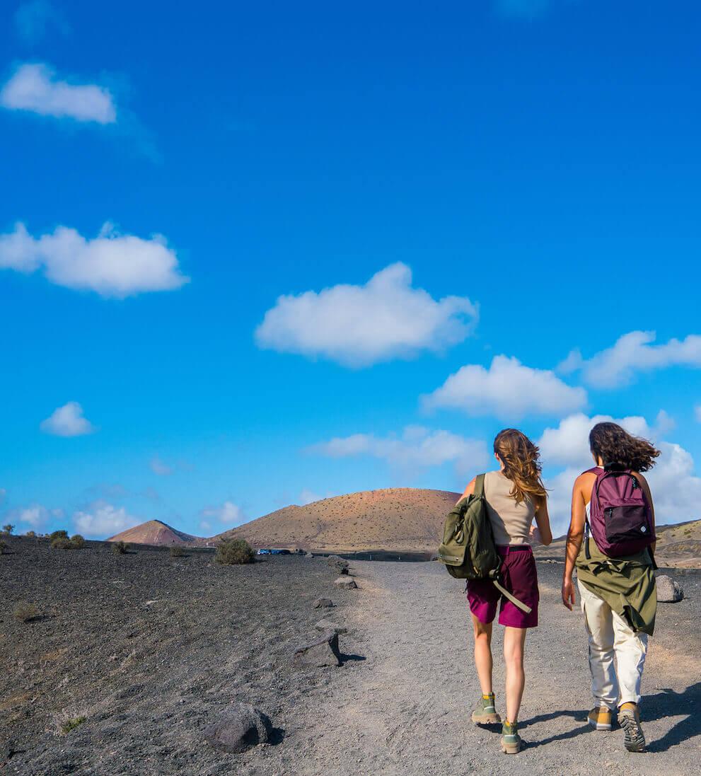 Due persone che camminano lungo un sentiero vulcanico nel Parco Nazionale di Timanfaya sotto un cielo blu.