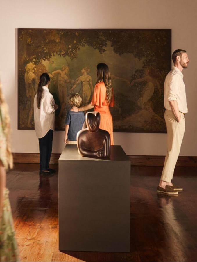 Gruppo in visita a una mostra d'arte in museo con quadri grandi e sculture in sala illuminata.